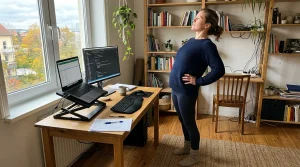 A remote worker performing a standing lumbar extension stretch at a home workstation with an ergonomic laptop setup