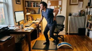 A desk worker standing up at the end of the day supporting his lower back with a floor back stretcher lying on an exercise mat
